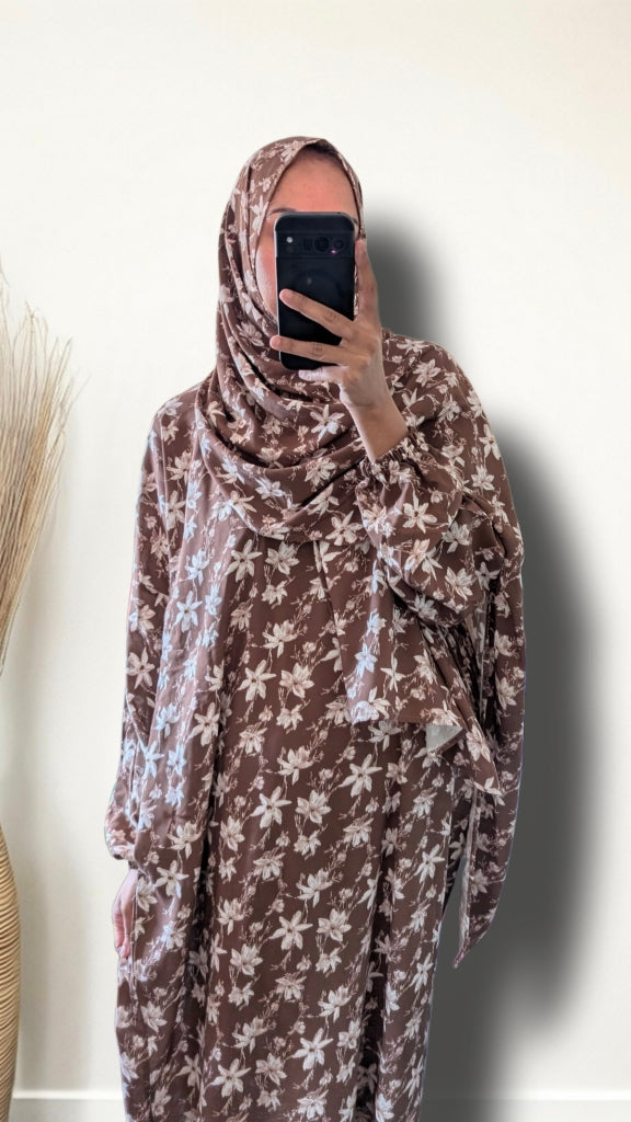 A person taking a mirror selfie wearing a brown and white floral print hijab and matching long-sleeve dress against a plain off-white wall.