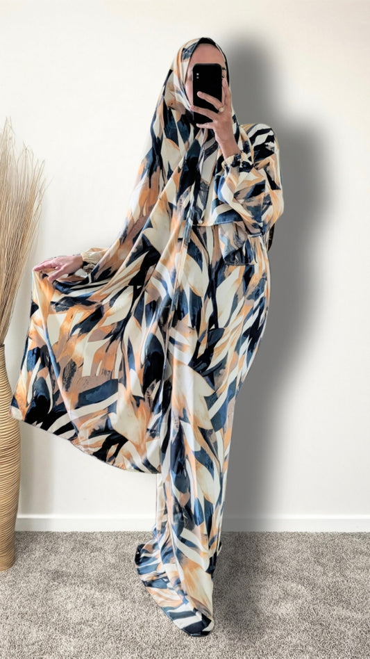 A woman wearing a flowing abstract-patterned kaftan dress in beige, navy blue, and orange, holding a black smartphone to take a mirror selfie against a plain white wall.
