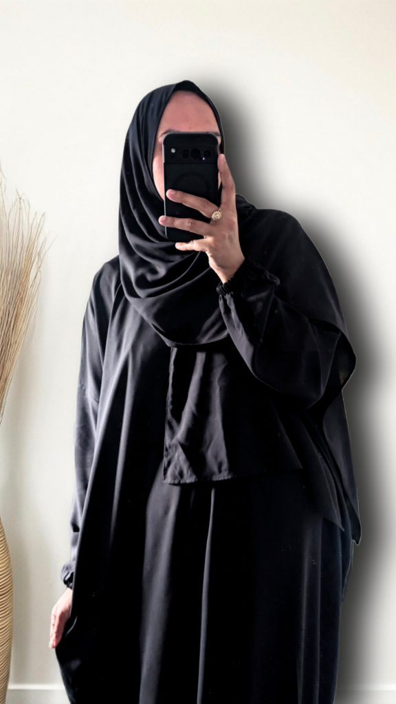 A person taking a mirror selfie wearing a full black modest outfit, including a black hijab and a long black abaya, holding a black smartphone to capture the fit. The background is a plain off-white wall with a dried decorative plant on the left.