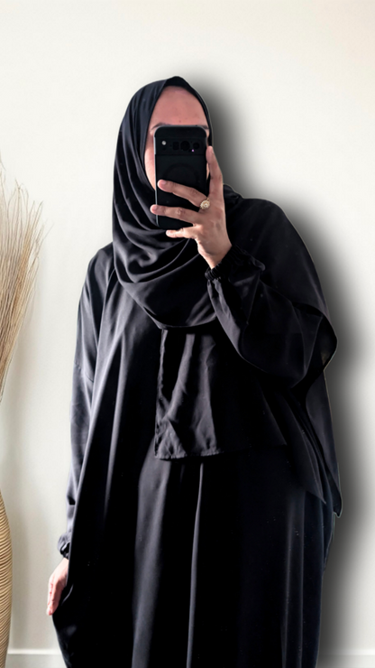 A person taking a mirror selfie wearing a full black modest outfit, including a black hijab and a long black abaya, holding a black smartphone to capture the fit. The background is a plain off-white wall with a dried decorative plant on the left.
