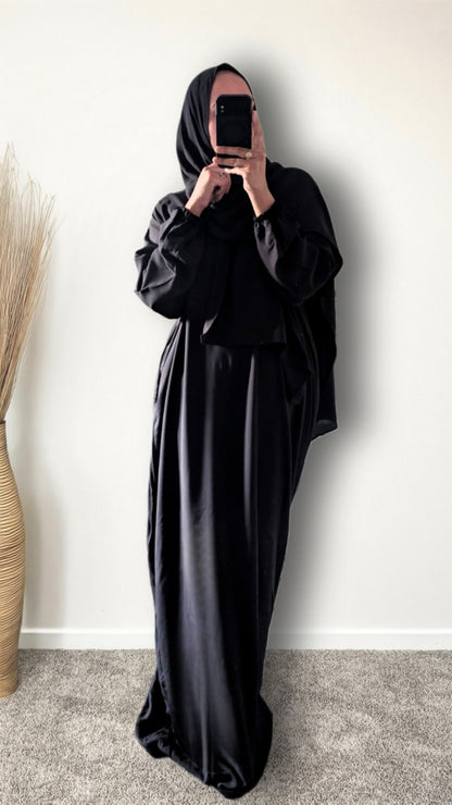 A person wearing a full black modest outfit with a long flowing abaya and matching head covering, taking a mirror selfie against a plain light wall.