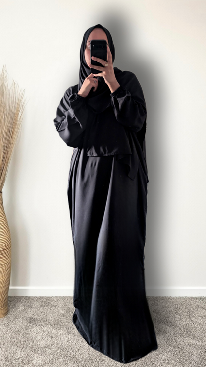 A woman wearing a black modest outfit with a long flowing abaya and matching hijab, taking a mirror selfie in an indoor setting with a plain wall and carpet.
