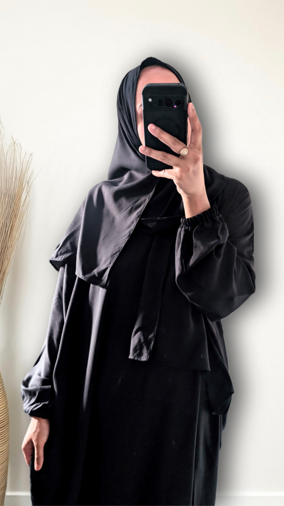 A person wearing a black abaya and matching hijab takes a mirror selfie against a plain off-white wall, holding a black smartphone.