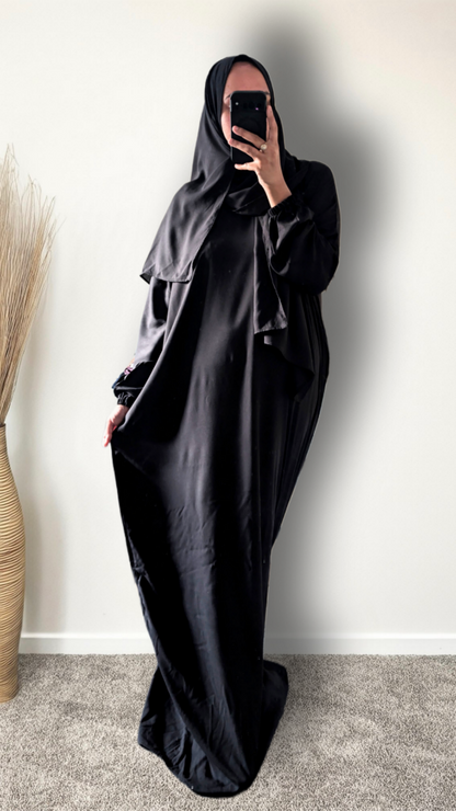 A person wearing a full black modest outfit with a headscarf and long draped garment, taking a mirror selfie indoors against a plain light wall.