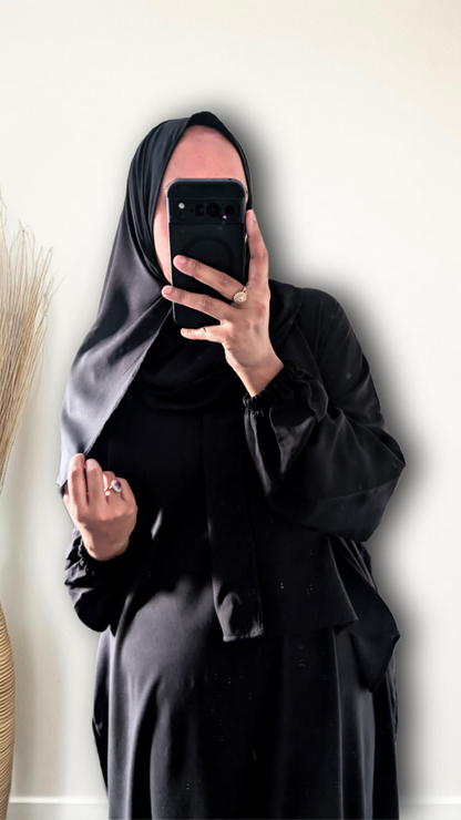 A person taking a mirror selfie wearing a full black modest outfit, including a black hijab and a long black abaya or robe, holding a black smartphone to block the face. The background is a plain off-white wall with a dried decorative plant on the left.