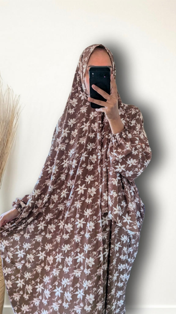 A person taking a mirror selfie wearing a brown and white floral print full-body garment with a matching head covering, holding a black smartphone in front of their face against a plain off-white wall.