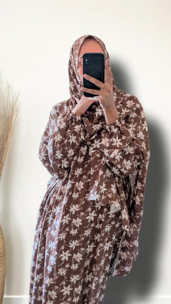 A person taking a mirror selfie wearing a brown and beige floral print hijab and a matching long-sleeve robe with a tie waist, holding a black smartphone.