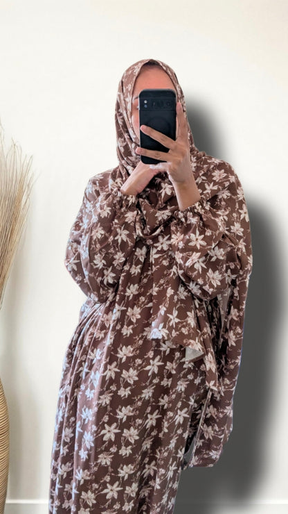A person taking a mirror selfie wearing a brown and beige floral print hijab and a matching long-sleeve robe with a tie waist, holding a black smartphone.
