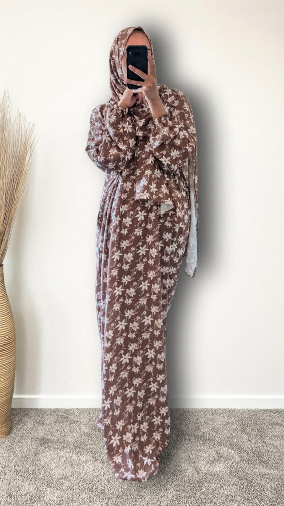 A woman wearing a brown and beige floral-patterned maxi abaya and matching hijab, taking a mirror selfie in an indoor setting.
