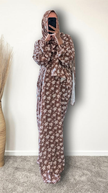 A woman wearing a brown and beige floral-patterned maxi abaya and matching hijab, taking a mirror selfie in an indoor setting.