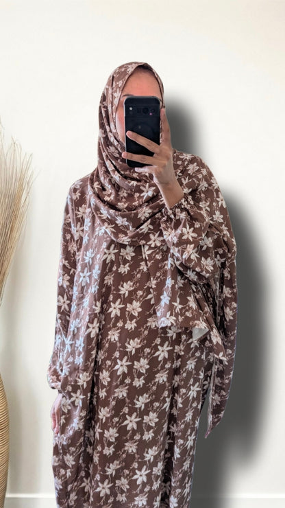 A person taking a mirror selfie wearing a brown and white floral print hijab and matching long-sleeve dress against a plain off-white wall.
