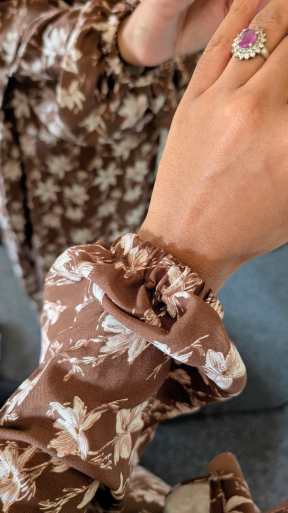 Close-up of a person's hand wearing a brown and beige floral print long-sleeve top, featuring a gathered ruffled cuff and a pink gemstone ring.