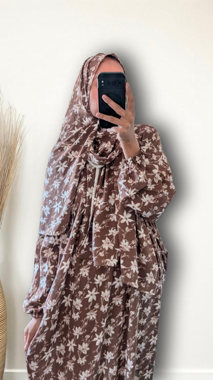 A person taking a mirror selfie wearing a brown and beige floral print modest dress with a matching headscarf, holding a black smartphone in front of their face against a plain off-white wall.