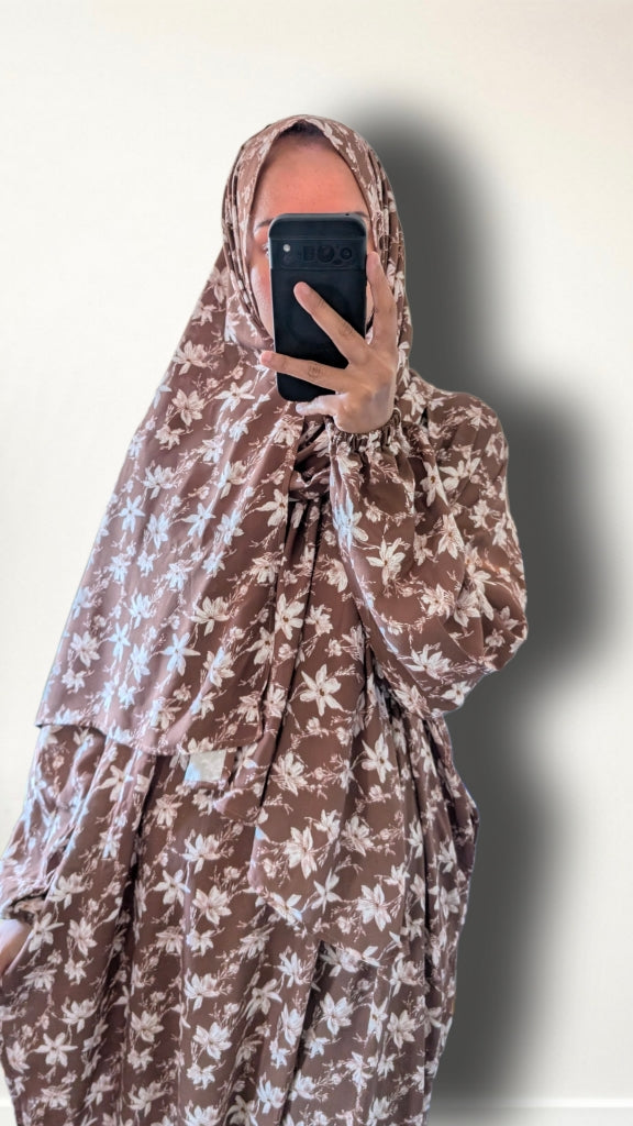 A person taking a mirror selfie wearing a brown and beige floral print abaya and matching headscarf, holding a black smartphone to block the face.