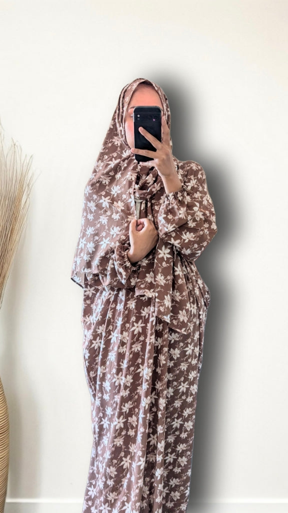 A woman wearing a brown and beige floral print hijab and matching long robe, holding a black smartphone to take a mirror selfie against a plain off-white wall.