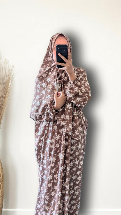 A woman wearing a brown and beige floral print hijab and matching long robe, holding a black smartphone to take a mirror selfie against a plain off-white wall.