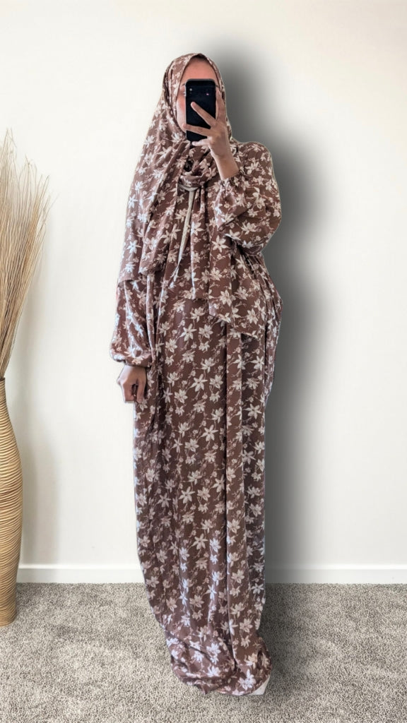 A woman wearing a long brown and beige floral print hijab and matching maxi dress, taking a mirror selfie in an indoor setting with a plain wall and a decorative vase.