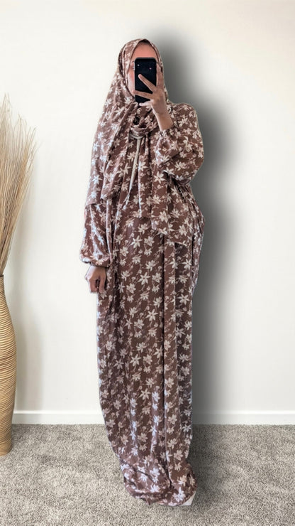 A woman wearing a long brown and beige floral print hijab and matching maxi dress, taking a mirror selfie in an indoor setting with a plain wall and a decorative vase.