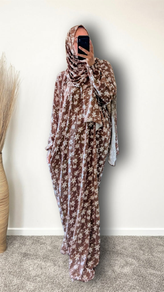 A woman wearing a brown and beige floral print hijab and a matching long kaftan dress, taking a mirror selfie in an indoor setting with a plain wall and a vase of dried grass.