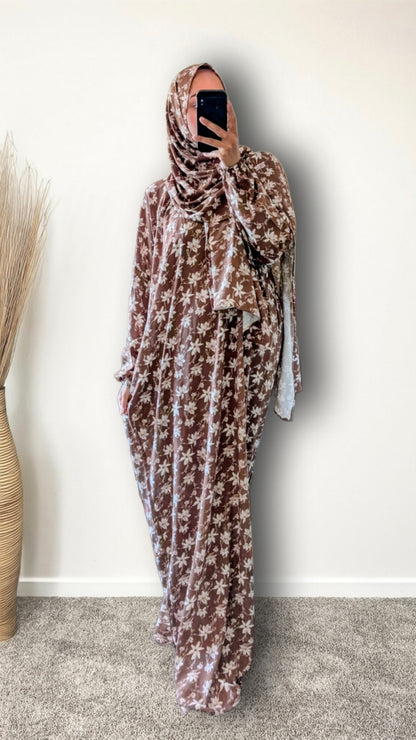 A woman wearing a brown and beige floral print hijab and a matching long kaftan dress, taking a mirror selfie in an indoor setting with a plain wall and a vase of dried grass.