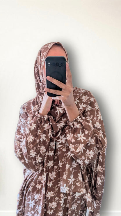 A person taking a mirror selfie wearing a brown and beige floral print hijab and matching long-sleeve dress against a plain off-white wall.