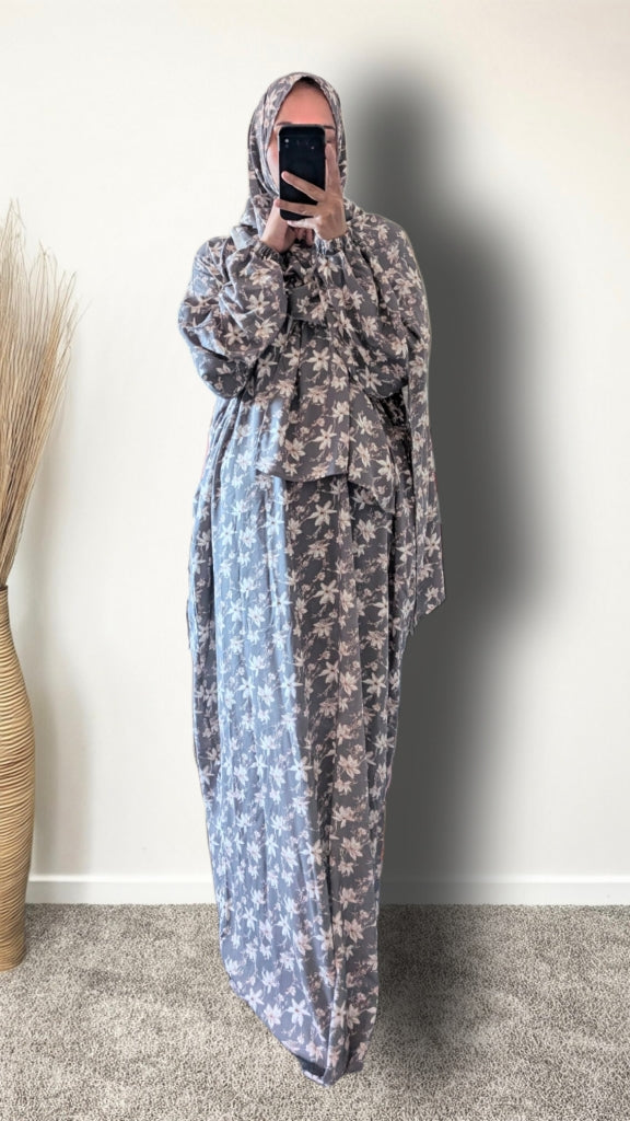 A person wearing a long, loose-fitting modest dress with a floral print in muted blue-gray and beige, paired with a matching headscarf, taking a mirror selfie indoors.