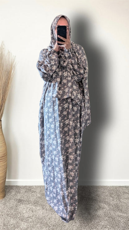 A person wearing a long, loose-fitting modest dress with a floral print in muted blue-gray and beige, paired with a matching headscarf, taking a mirror selfie indoors.