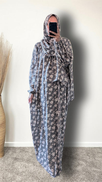 A woman wearing a long, loose-fitting modest dress with a gray and white floral pattern, paired with a matching hijab, taking a mirror selfie in an indoor setting.