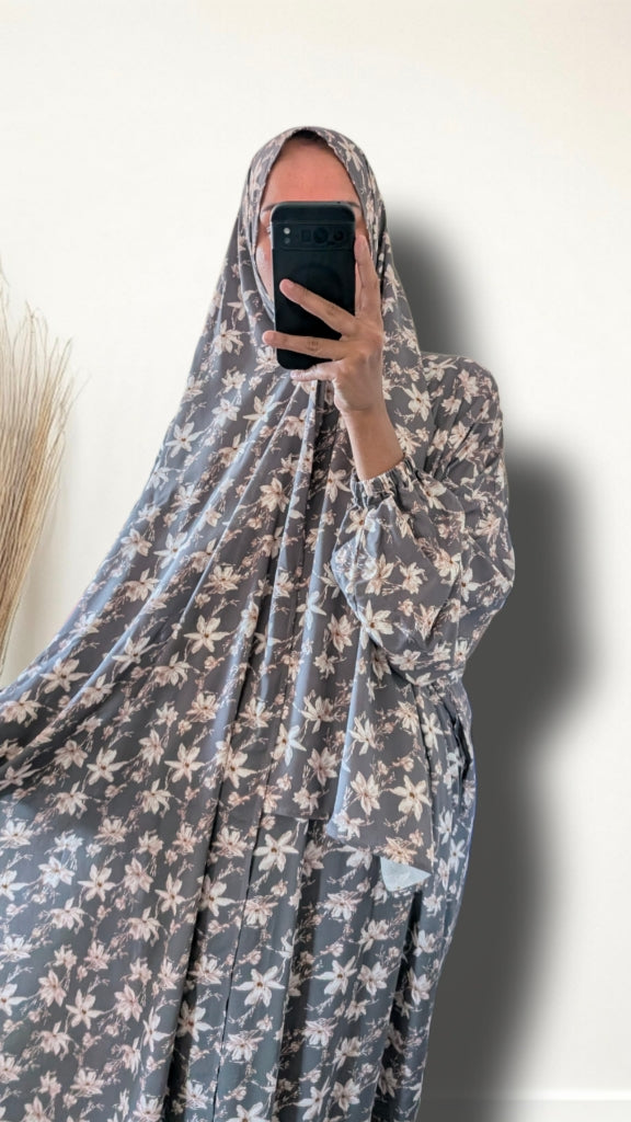 A person taking a mirror selfie wearing a full-body modest garment with a gray base and white floral pattern, holding a black smartphone in front of their face against a plain light wall.