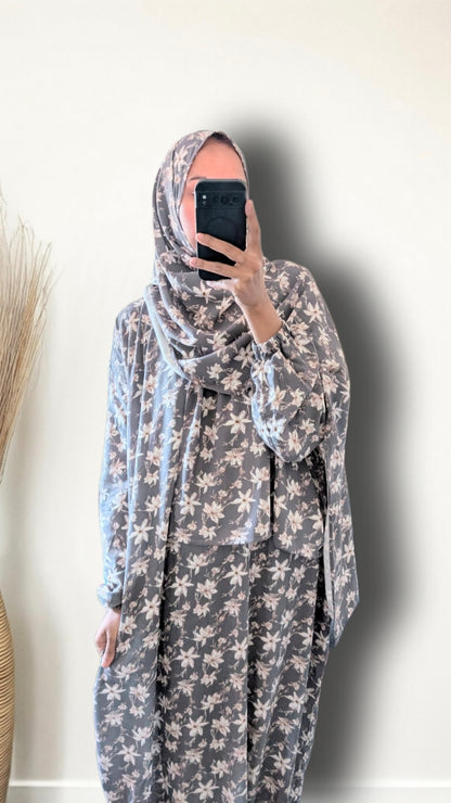 A woman wearing a gray and white floral print hijab and matching long-sleeve abaya, taking a mirror selfie against a plain off-white wall.
