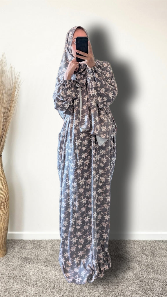 A woman wearing a long, loose-fitting maxi dress with a grey and white floral pattern, paired with a matching headscarf, taking a mirror selfie in an indoor setting.