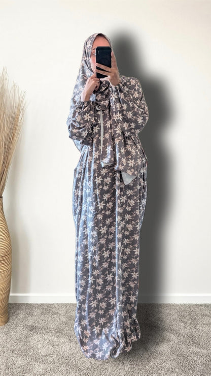 A woman wearing a long, loose-fitting maxi dress with a grey and white floral pattern, paired with a matching headscarf, taking a mirror selfie in an indoor setting.