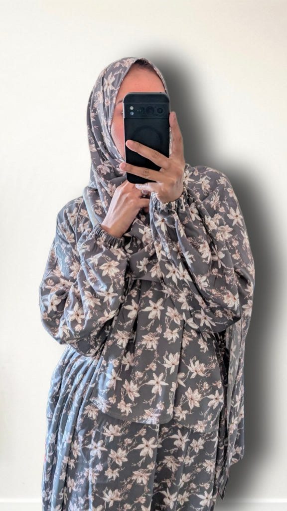 A person taking a mirror selfie wearing a gray and beige floral print hijab and matching long-sleeve garment against a plain off-white wall.
