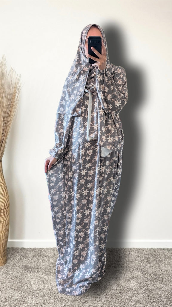 A woman wearing a long, flowing maxi dress with a matching head covering, featuring a light blue and white floral pattern. She is taking a mirror selfie in an indoor setting with a plain wall and a carpeted floor.