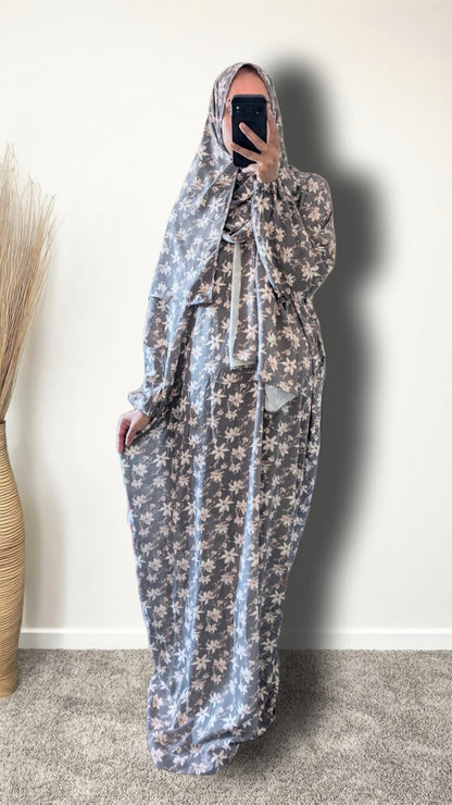 A woman wearing a long, flowing maxi dress with a matching head covering, featuring a light blue and white floral pattern. She is taking a mirror selfie in an indoor setting with a plain wall and a carpeted floor.