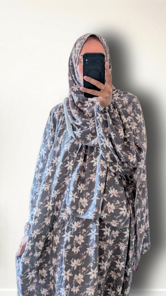 A person taking a mirror selfie wearing a loose, long-sleeved floral hijab and shawl in a gray and beige pattern against a plain off-white wall.
