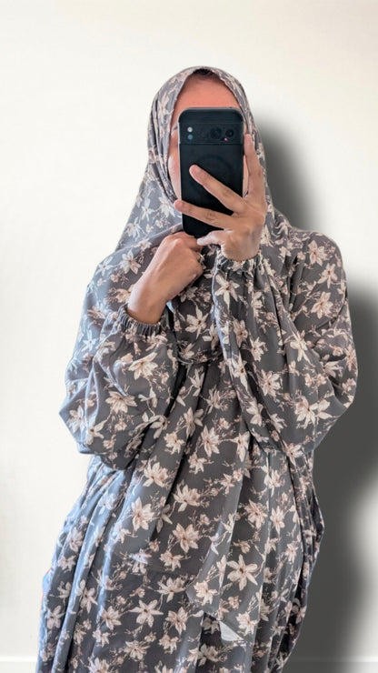 A person wearing a long-sleeved gray hijab and matching modest dress with a light beige floral pattern, holding a black smartphone to take a mirror selfie against a plain off-white wall.