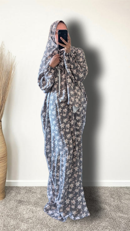 A woman wearing a long, loose-fitting maxi dress with a matching head covering, featuring a gray and white floral pattern. She is holding a black smartphone to take a mirror selfie in an indoor setting with a plain off-white wall and a beige carpet.