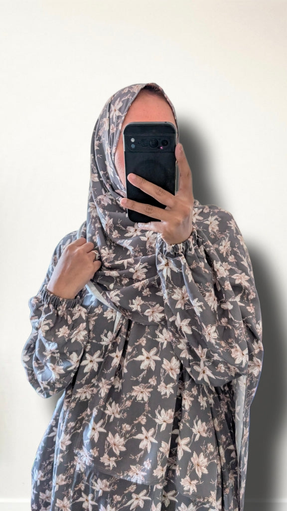 A person taking a mirror selfie wearing a long-sleeved, full-body floral print garment in gray and beige, with a matching head covering. The background is a plain off-white wall.