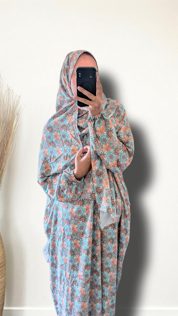 A person taking a mirror selfie wearing a loose, long-sleeved abaya dress with a blue and peach floral pattern, paired with a matching patterned hijab, set against a plain off-white wall.