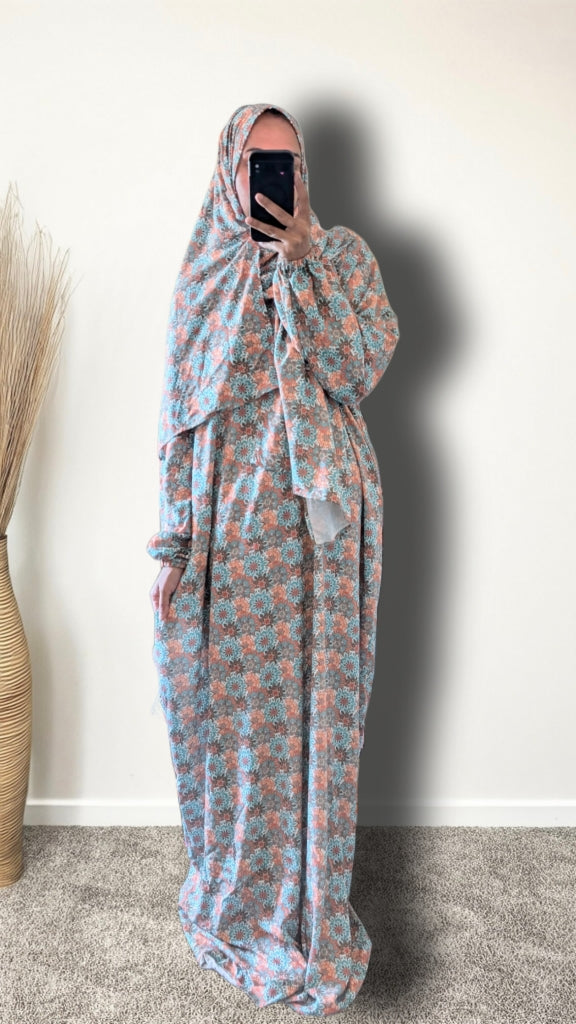 A person wearing a full-length modest outfit with a matching headscarf and long sleeves, featuring a light blue and peach floral pattern, taking a mirror selfie against a plain wall.
