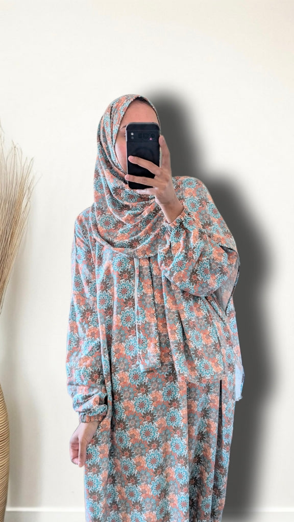 A woman wearing a matching floral hijab and long-sleeve abaya with a matching skirt, taking a mirror selfie against a plain off-white wall.