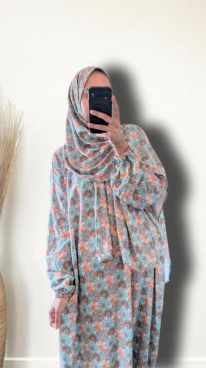 A woman wearing a matching floral hijab and long-sleeve abaya with a matching skirt, taking a mirror selfie against a plain off-white wall.