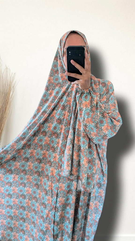 A person taking a mirror selfie wearing a full-body modest garment with a blue and peach floral pattern, set against a plain off-white wall.