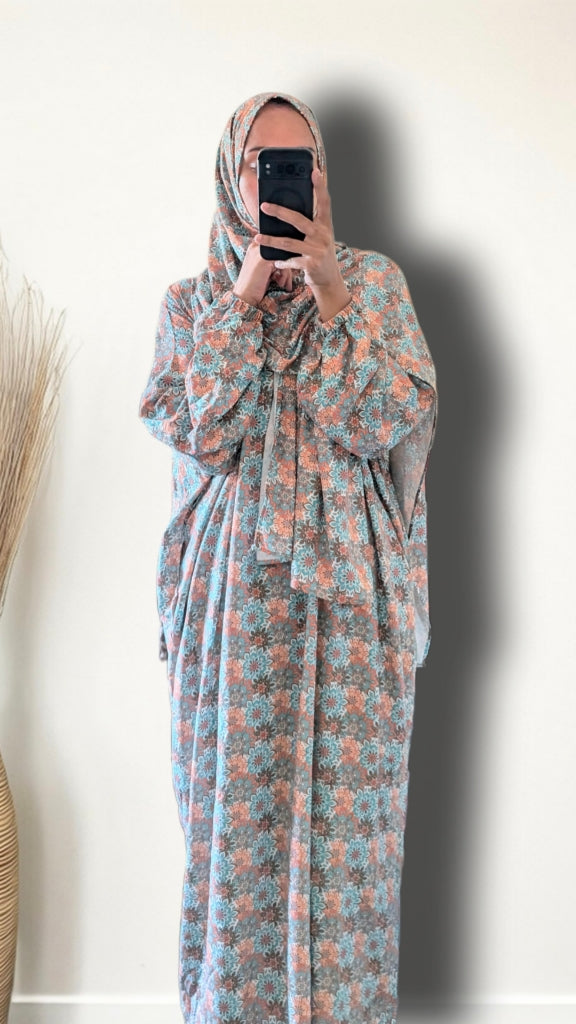 A woman wearing a long-sleeved, floor-length modest dress with a floral pattern in dusty blue and terracotta orange, paired with a matching patterned hijab, taking a mirror selfie against a plain off-white wall.