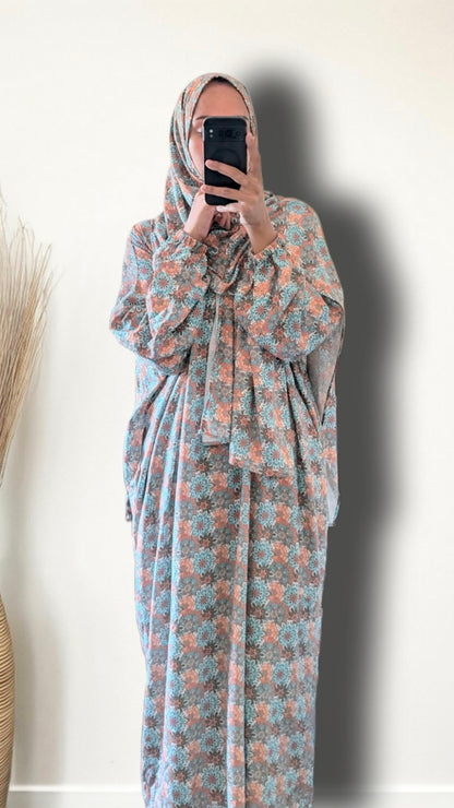 A woman wearing a long-sleeved, floor-length modest dress with a floral pattern in dusty blue and terracotta orange, paired with a matching patterned hijab, taking a mirror selfie against a plain off-white wall.