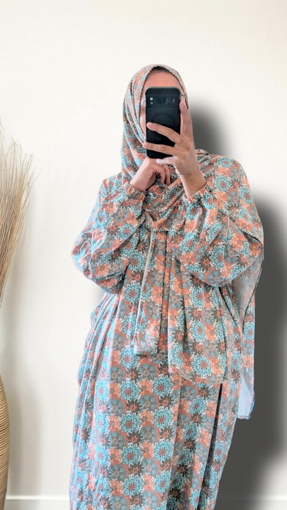 A woman takes a mirror selfie wearing a long-sleeved maxi abaya dress with a blue and peach floral pattern, paired with a matching patterned hijab.