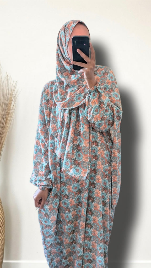 A woman takes a mirror selfie wearing a long-sleeved, loose-fitting modest dress with a matching hijab. The fabric features an all-over floral pattern in dusty pink, light blue, and brown.