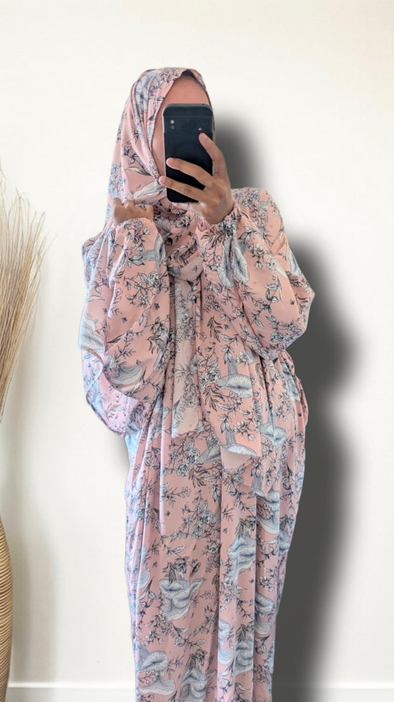 A person takes a mirror selfie wearing a long-sleeved, full-length modest dress or abaya in a dusty pink color with a blue and gray floral pattern, paired with a matching patterned hijab. The background is a plain off-white wall with a shadow cast to the right.