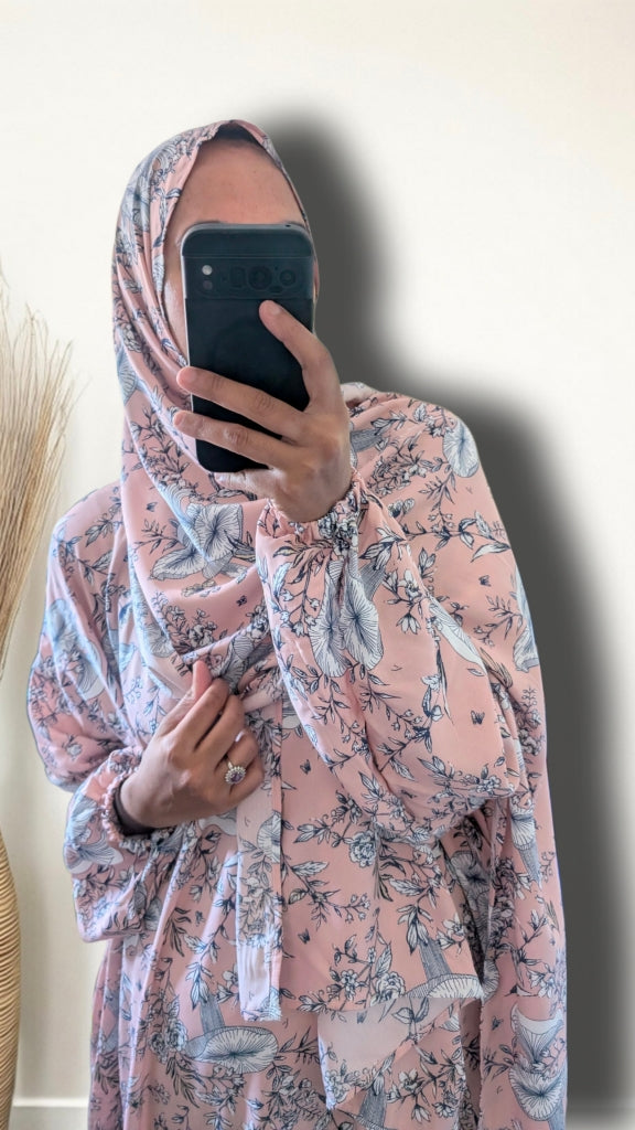 A person taking a mirror selfie wearing a light pink hijab and a matching long-sleeve tunic with a blue and gray toile-style floral print. The background is a plain off-white wall with a dark shadow cast to the right.